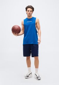 Νέος άνδρας που φορά μπλε αμάνικο πουκάμισο και σορτς, κρατώντας μια μπασκέτα στο αριστερό του χέρι, στέκεται μπροστά σε ένα απλό φωτεινό φόντο.