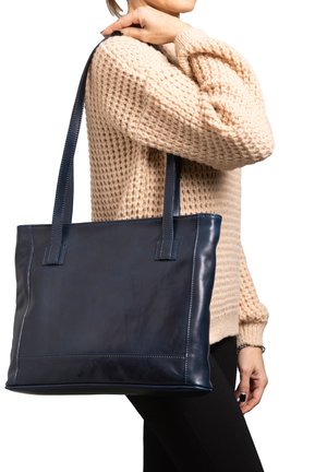 Person wearing beige knit sweater and black pants carries large navy blue leather tote bag over shoulder.