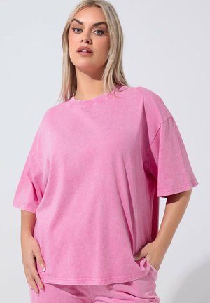 Blonde vrouw die een losse roze shirt met korte mouwen en bijpassende broek draagt, staand met handen in de zakken tegen een effen achtergrond.