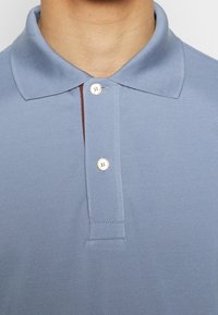 Gros plan sur une personne portant un polo bleu clair avec une patte à deux boutons et une doublure intérieure marron visible.