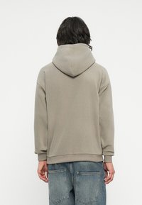 Grå hettegenser med en myk tekstur, avslappet passform, ribbede mansjetter og hem, brukt over denimbukser, sett fra baksiden.