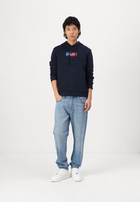 Navy hoodie with a front pocket and drawstring, featuring a red and white logo panel. Worn with light blue jeans and white sneakers.