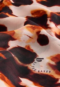 Zijdeachtige stof in warme tinten van oranje en bruin met abstracte patronen. Bevat een logo in donkere letters afgedrukt nabij de onderste rand.