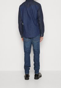 Camisa de mezclilla oscura con hombros más claros, mangas largas y un diseño de yugo curvado; combinada con jeans azules medianos y zapatos negros.