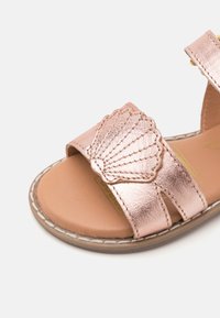 Roséguld metalliska sandaler med en snäcka-formad dekoration, som har två remmar och en beige sula med vit sömdekoration.