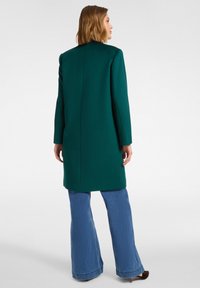 Grüner Wollmantel mit geradem Schnitt, langen Ärmeln und Gehschlitz hinten, kombiniert mit weitem Denim-Jeans. Glatte Textur, strukturierte Silhouette.