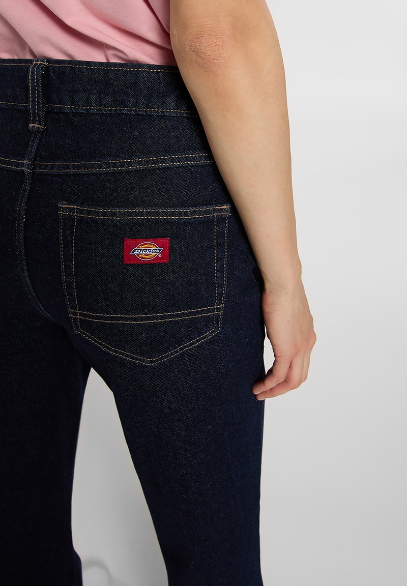 Dark blue denim jeans with a back pocket showcasing a red and yellow branded tag, and contrasting yellow stitching along the seams.