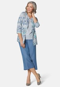 Lichtblauwe geprinte cardigan over een lichtgrijze top, gecombineerd met blauwe cropped broeken. De cardigan heeft een bloemenpatroon en driekwart mouwen.