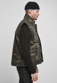 Zöld és barna szövetből készült camo mellény magas gallérral, ujjatlan kialakítással, elülső zsebekkel és patentos záródással. Fekete, texturált pulóver fölött viselve.