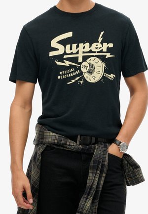 Homme portant un t-shirt graphique noir "Super Dry" avec des éclairs, une chemise à carreaux nouée autour de la taille, un pantalon noir et une montre-bracelet.