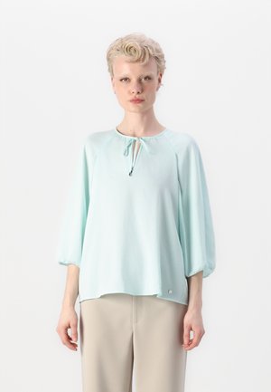 Helles mintgrünes Blusenshirt mit rundem Halsauschnitt, Bindedetail an der Vorderseite und drei Viertel Puffärmeln. Besitzt einen Logoakzent am Saum.