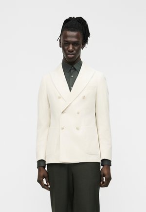 Mand med mørk hud og dreadlocks iført en dobbeltradet cremefarvet blazer over en mørkegrøn skjorte med knapper og matchende bukser, stående mod en hvid baggrund.