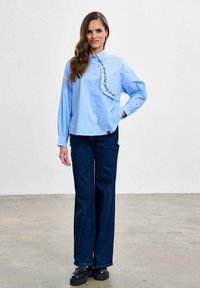 Femeie purtând o bluză albastru deschis cu detalii text, blugi albaștri închiși și pantofi platformă negri, stând pe un podea de beton în fața unui perete alb.