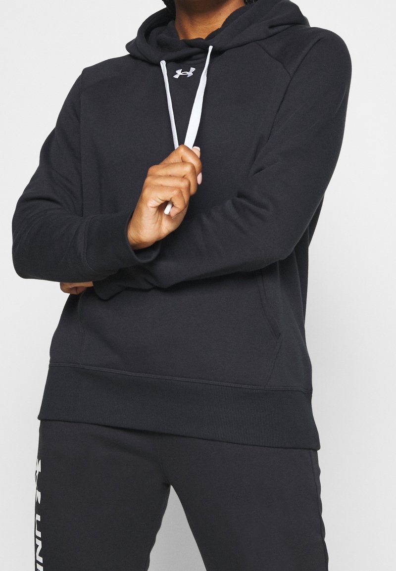 Personne portant un sweat à capuche noir Under Armour et un pantalon de survêtement noir assorti, tenant un cordon de capuche blanc avec les bras croisés.