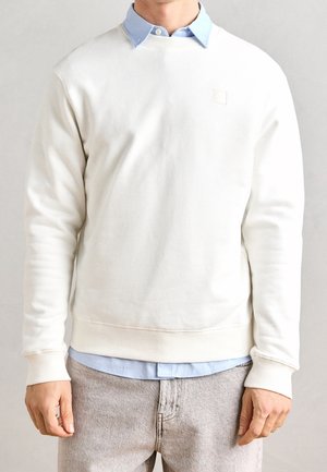 Mand iført hvid crewneck sweatshirt over lyseblå skjorte med krave og lysegrå jeans, stående med hænderne afslappet ned langs siderne.