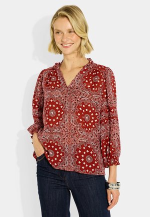 Mujer sonriente con una blusa roja con estampado y mangas abullonadas y vaqueros oscuros, de pie con una mano en el bolsillo frente a un fondo liso.