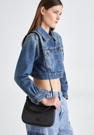 Kobieta w krótkiej kurtce dżinsowej i jeansach w stylu acid wash, niosąca czarną torebkę na ramię z teksturowanym wykończeniem i detalem z logo.