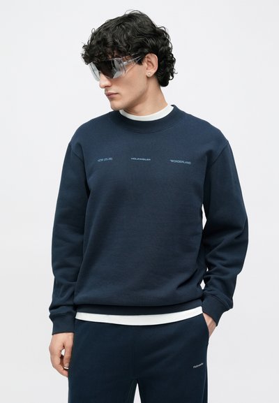 Temno modra jopa s krožnim izrezom, dolgimi rokavi in kontrastnim belim napisom. Lahka tkanina, sproščen kroj, v kombinaciji z usklajenimi hlačami.