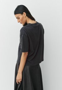 Zwarte katoenen T-shirt met korte mouwen, ronde halslijn, losse pasvorm, met strassteentjes accent op de linkerschouder en zijkant.