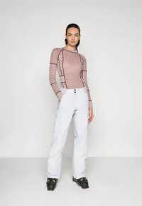 Nő áll, testhez álló mintás, hosszú ujjú felsőt, fehér, magas derekú hónadrágot és fekete sícipőt viselve egy sima háttér előtt.