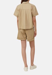 Beżowa koszula z krótkim rękawem w luźnym kroju z plisą z tyłu, w zestawieniu z dopasowanymi szortami z podwiniętymi mankietami. Uzupełniona jasnymi sneakersami.