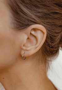 Pendiente de aro plateado, pequeño y Delgado, cuelga del lóbulo de una oreja. El diseño es suave y pulido con un brillo sutil.