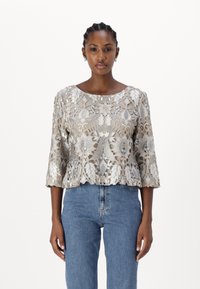 Beige bestickte Bluse mit Dreiviertelärmel, Overlay-Muster aus Blättern und Blumen, gewellter Saum, kombiniert mit blauen Jeans.