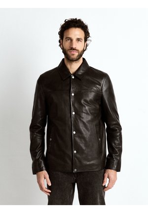 Mann trägt eine schwarze Lederjacke mit Knöpfen, Kragen und langen Ärmeln, steht vor einem einfarbigen weißen Hintergrund.