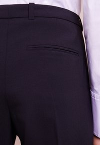 Marineblauwe pantalon met een gladde textuur, een slimfit pasvorm en een enkele achterzak. De tailleband heeft een gepolijste zwarte knoopsluiting.