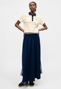 Femme debout, les mains sur les hanches, portant un polo en maille crème et une longue jupe bleu marine à rayures blanches avec des baskets noires.