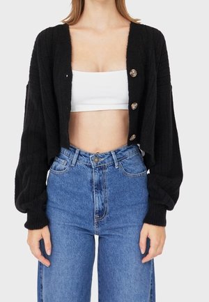 Kobieta ubrana w czarny krótki kardigan na guziki na białym krótki topie i niebieskich jeansach z wysokim stanem, pokazana od szyi do bioder.