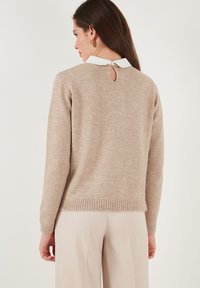 Donna che indossa un maglione beige a maniche lunghe con colletto bianco e orecchini a cerchio dorati, vista di spalle su uno sfondo neutro.