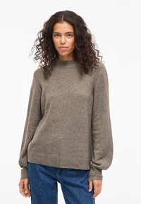 Ženska s kodrastimi temnimi lasmi, oblečena v rjav pleten pulover in modre kavbojke, stoji pred enobarvno belo ozadjem.