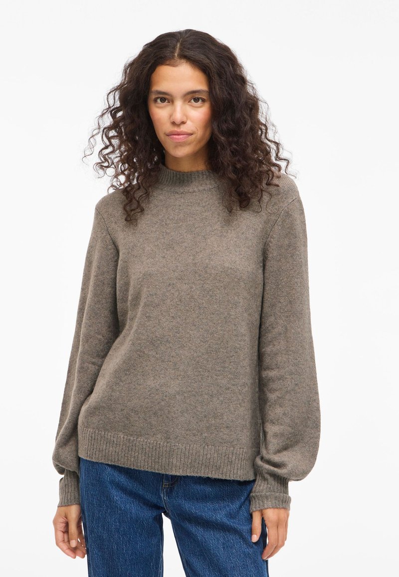 Ženska s kodrastimi temnimi lasmi, oblečena v rjav pleten pulover in modre kavbojke, stoji pred enobarvno belo ozadjem.