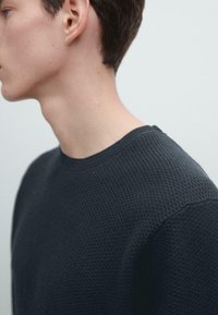 Maglione blu navy con trama a motivo waffle e bordi a coste, con scollatura tonda, visto di lato su sfondo neutro.