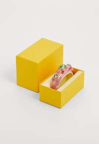 Pink resin bangle with three green gemstones in gold settings, resting in a yellow box with a light-colored interior.
