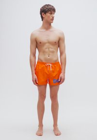 Πορτοκαλί μαγιό βερμούδα με λευκό κορδόνι, που διαθέτει μπλε λογότυπο "DS" στο αριστερό πόδι. Ελαφρύ, απαλό ύφασμα, μήκος έως τη μέση του μηρού.