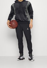 Schwarze Velours-Hoodie mit grauen Einsätzen, Kängurutasche und Logo, kombiniert mit schwarzen Jogginghosen mit weißem Logo, während ein Basketball gehalten wird.