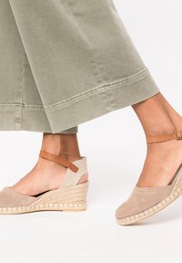 Cuñas de espadrille beige con parte superior texturizada, puntera de ante y suela de yute trenzado. Correa ajustable para el tobillo en tono marrón y diseño de talón abierto.