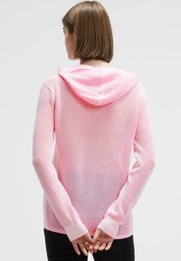 Frau mit kurzen braunen Haaren, die einen hellrosa luftigen Strick-Hoodie trägt, steht mit hinter dem Rücken verschränkten Händen vor einem schlichten Hintergrund.