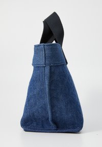 Denim soma ar ģeometrisku trīsstūra formu, tumši zila krāsa un melns auduma rokturis. Iespējama redzama šuvju apdare un strukturēta augšdaļa.