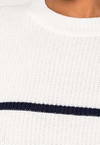 Weißer Strickpullover mit einem horizontalen marineblauen Streifen über der Brust. Der runde Ausschnitt und das strukturierte Material sorgen für ein angenehmes Aussehen.