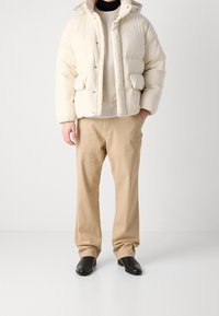 Puffjakke i creme med hætte, med trykknapper og sidelommer. Sammen med beige bukser og sorte sko.