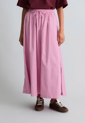 Frau trägt einen rosa plissierten Maxirock mit vertikalen schwarzen Streifen, braune Sneaker mit weißen Schnürsenkeln und ein dunkelrotes Oberteil.