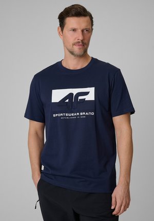 Mężczyzna w granatowej koszulce z krótkim rękawem z logo „4F Sportswear Brand Established in 1995”, stojący na jednolitym szarym tle.