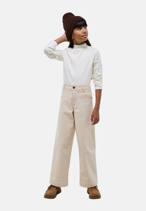 Pantalons larges beiges, top à manches longues blanc côtelé, et bonnet en tricot marron. Chaussures brunes avec des détails texturés ; modèle posé debout.