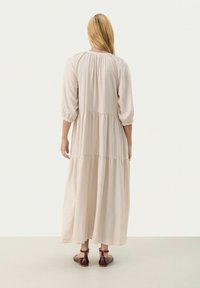 Rochie lungă, bej, cu mânici bufante trei sferturi, design în straturi și o siluetă fluidă; asortată cu sandale cu curea de culoare maro închis.