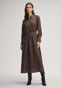 Lungo vestito marrone con motivo in pelle di coccodrillo, scollo colletto, chiusura con bottoni e una cintura in vita, abbinato a stivali neri fino al ginocchio.