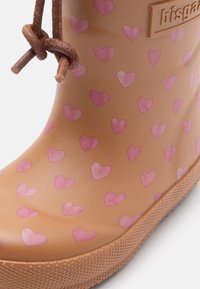 Botas de lluvia de goma marrón con patrones de corazones rosas, suela texturizada y detalles de cordones marrones. Presenta una placa con el logo en el lado exterior.