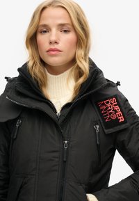 Sort pufferjakke med lynlås, høj krave og et mærke med "SUPER DRY MNTN" i rød. Har struktureret stof og flere lommer.